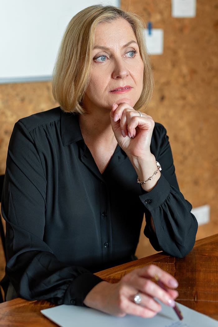 Mature businesswoman in black attire thinking at a wooden desk in a modern office.